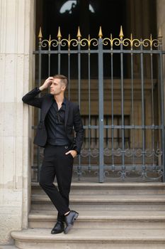 Young man standing on concrete stairs near metal gate and leaning on building. Concept of fashion male model. Boy wearing black suit.