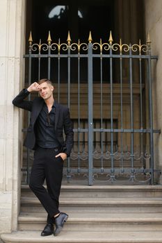 Young european man standing near metal gate and leaning on building. Concept of fashion male model. Boy wearing black suit.