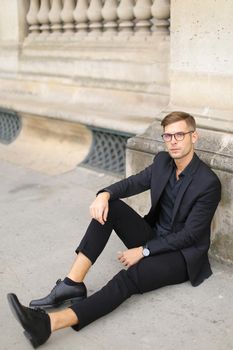 Young european man sitting on sidewalk ground and wearing black suit, leaning on concrete banister. Concept of walking in cty and male fashionable model.