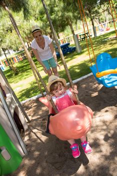 young mother and her little daughter smiling together while swinging at the playground in the park