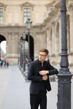 Young man walking in Paris and leaning on lantern, weaaring black suit. Concept of walking in city and male fashion.