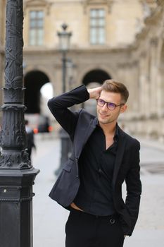 Young handsome man in glasses walking in city and wearing black suit, leaning on lantern. Concept of strolling and male fashion model, urban photo session.