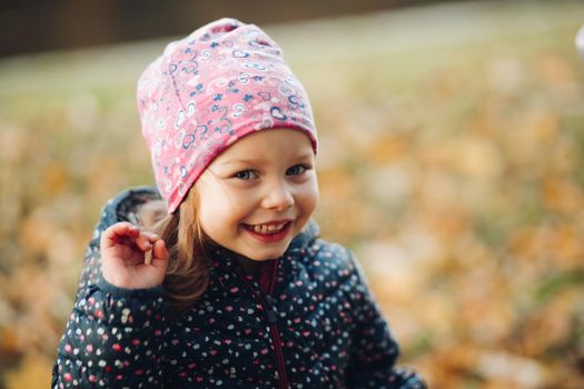 Stock photo of a cute little young girl wearing pink printed hat and dark printed winter jacket smiling at camera and waving hand. Standing against blurred autumnal background.