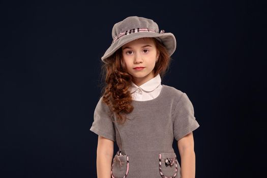 Studio shot of a lovely little child with a long, curly hair looking at the camera. Charming girl in a combo dress and a gray hat is posing on a black background. Sincere emotions concept.