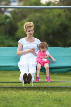 A beautiful young mother and her happy little daughter enjoying free time while swinging on bench at backyard