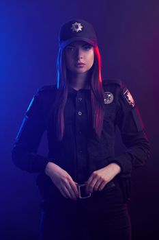 Redheaded girl police officer in a dark uniform and a cap, with bright make-up, is holding her belt and looking at the camera against a black background with red and blue backlighting.