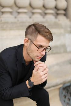 Young european man sitting on sidewalk and leaning on concrete railing of building, wearing black suit and glasses. Concept of walkig in city, urban photo session and male person model.