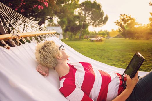 young woman using a tablet computer while relaxing on hammock in a peaceful garden during holiday