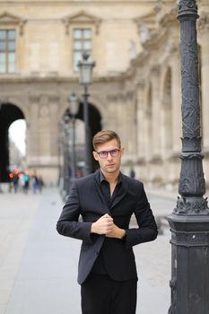 Young handsome man walking in Paris and leaning on lantern, weaaring black suit. Concept of walking in city and male fashion.