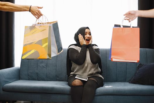 Modern arab girl in hijab at home, doing shopping online, while sitting on couch in living room. Two hand with packages. Concept of easy online shopping.