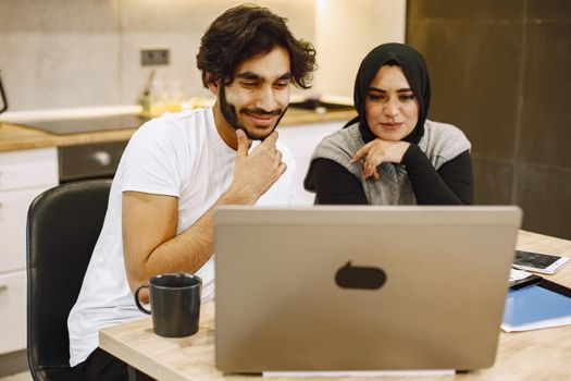 Beautiful young couple using a laptop, writing in a notebook, sitting in a kitchen at home. Arab girl wearing black hidjab.