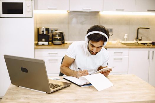 Arabic man watching online webinar, sitting in a kitchen with computer, enjoying distance learning.