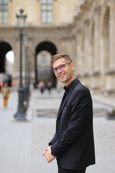 Young man standing near building in city and wearing black suit, lanterns in background. Concept of male fashion model and urban photo session.