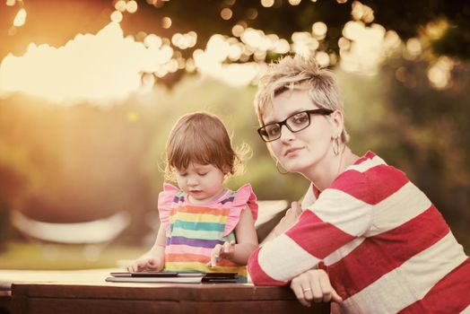 Happy mother and her little daughter enjoying free time using tablet computer while relaxing  on holiday home garden during sunny day