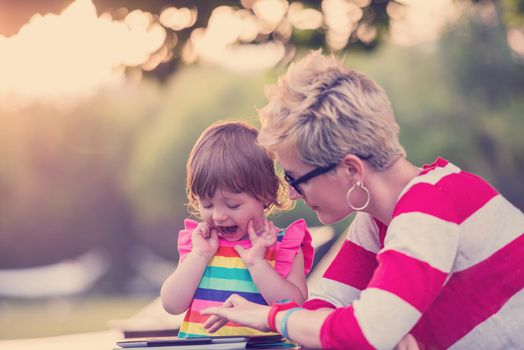 Happy mother and her little daughter enjoying free time using tablet computer while relaxing  on holiday home garden during sunny day