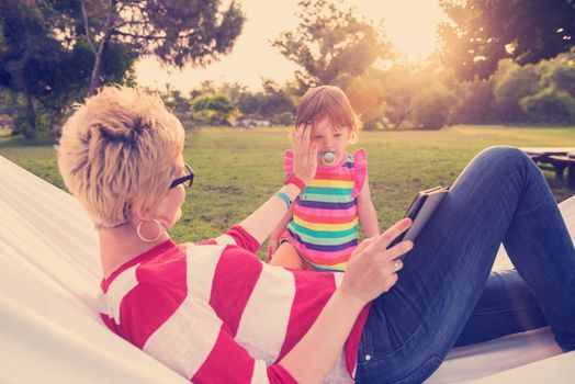 Happy mother and her little daughter enjoying free time using tablet computer while relaxing in a hammock during sunny day on holiday home garden