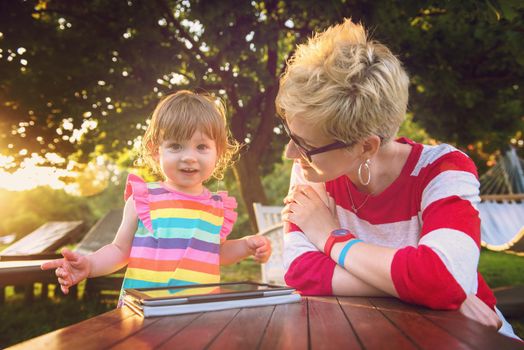 Happy mother and her little daughter enjoying free time using tablet computer while relaxing  on holiday home garden during sunny day