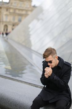 Young man in sunglasses wearing black jacket sitting near glass Louvre Pyramid in Paris and smoking cigarette, France. Concept of male fashion model and urban photo session.