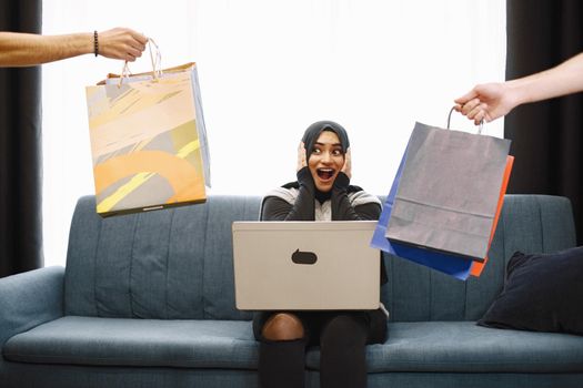 Modern arab girl in hijab using laptop at home, doing shopping online, while sitting on couch in living room. Two hand with packages. Concept of easy online shopping.