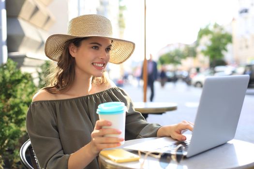 Happy nice woman working on laptop in street cafe, holding paper cup