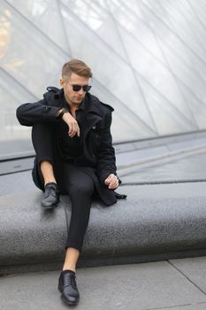 Young european man in sunglasses wearing black jacket sitting near glass Louvre Pyramid in Paris and smoking cigarette, France. Concept of male fashion model and urban photo session.