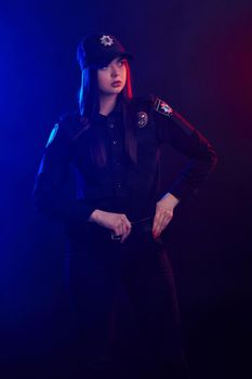 Ginger female police officer in a uniform and a cap, with bright make-up, is touching her belt and looking away against a black background with red and blue backlighting. Defender of citizens is ready to enforce a law and stop a crime.