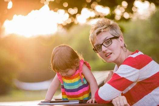 Happy mother and her little daughter enjoying free time using tablet computer while relaxing  on holiday home garden during sunny day