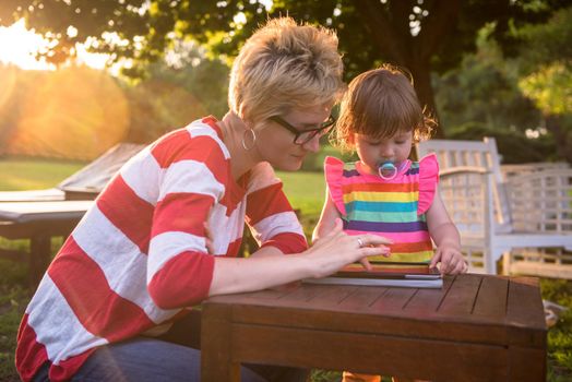 Happy mother and her little daughter enjoying free time using tablet computer while relaxing  on holiday home garden during sunny day