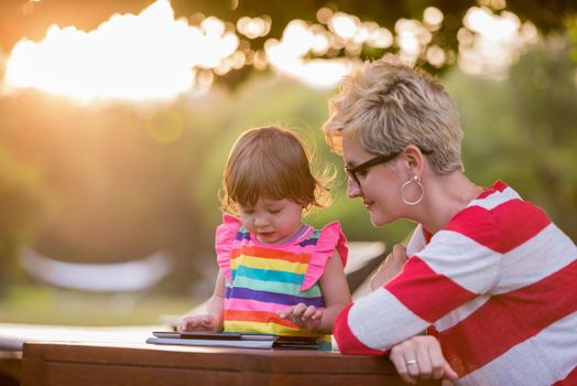 Happy mother and her little daughter enjoying free time using tablet computer while relaxing  on holiday home garden during sunny day