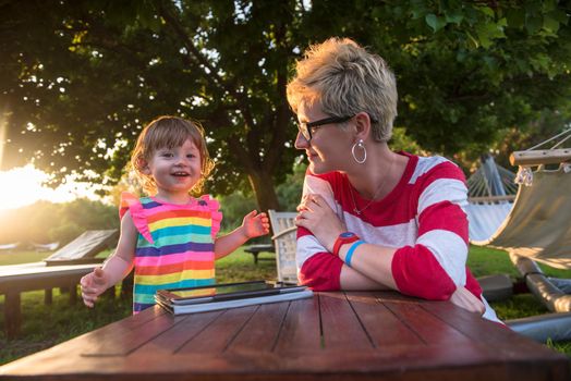 Happy mother and her little daughter enjoying free time using tablet computer while relaxing  on holiday home garden during sunny day