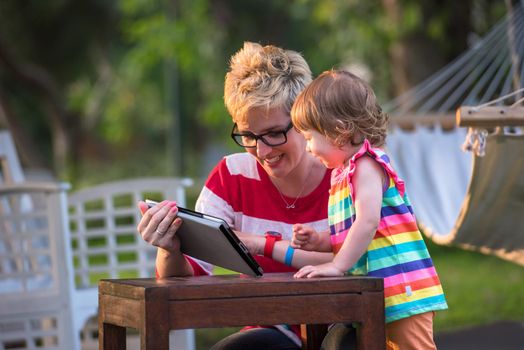 Happy mother and her little daughter enjoying free time using tablet computer while relaxing  on holiday home garden during sunny day