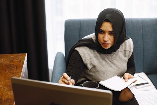 Modern arab girl in hijab using laptop at home, working remotely while sitting on couch in living room