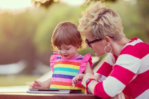 Happy mother and her little daughter enjoying free time using tablet computer while relaxing  on holiday home garden during sunny day