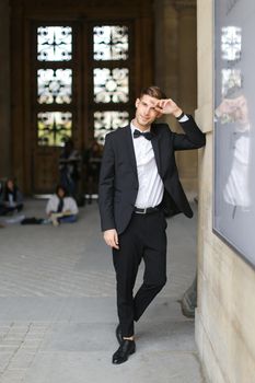 Young handsome man standing and leaning on building, wearing black suit and bow tie, window and sitting people in background. Concept of fashionable businessman and male model.