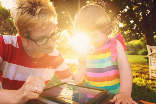 Happy mother and her little daughter enjoying free time using tablet computer while relaxing  on holiday home garden during sunny day