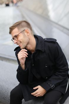 Young handsome man wearing black jacket sitting near glass Louvre Pyramid in Paris, France. Concept of male fashion model and urban photo session.