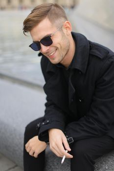 Young smiling man in sunglasses wearing black jacket sitting near glass Louvre Pyramid in Paris and smoking cigarette, France. Concept of male fashion model and urban photo session.