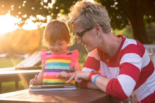 Happy mother and her little daughter enjoying free time using tablet computer while relaxing  on holiday home garden during sunny day