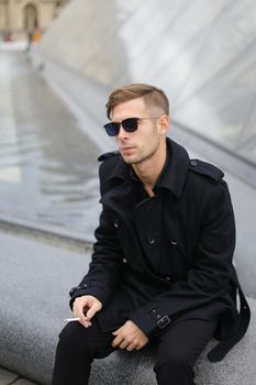 Young handsome man in sunglasses wearing black jacket sitting near glass Louvre Pyramid in Paris and smoking cigarette, France. Concept of male fashion model and urban photo session.