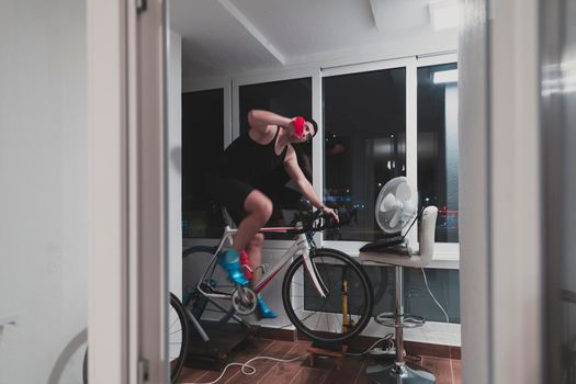 Man cycling on the machine trainer he is exercising in the home at night. Playing online bike racing game during coronavirus covid19 lockdown. New normal concept.