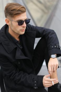 Young fashionable man in sunglasses wearing black jacket sitting near glass Louvre Pyramid in Paris and smoking cigarette, France. Concept of male fashion model and urban photo session.