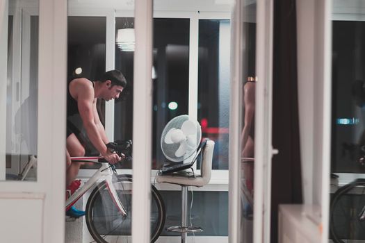 Man cycling on the machine trainer he is exercising in the home at night. Playing online bike racing game during coronavirus covid19 lockdown. New normal concept.