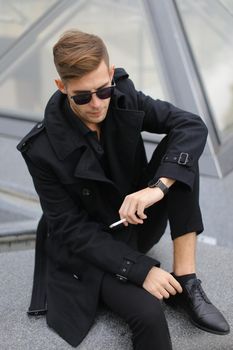 Young stylish man in sunglasses wearing black jacket sitting near glass Louvre Pyramid in Paris and smoking cigarette, France. Concept of male fashion model and urban photo session.