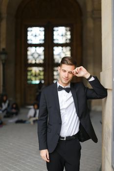 Young groom standing and leaning on building, wearing black suit and bow tie, window and sitting people in background. Concept of fashionable businessman and male model.