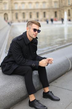 Young smiling man in sunglasses wearing black clothes sitting near glass Louvre Pyramid in Paris and smoking cigarette, France. Concept of male fashion model and urban photo session.