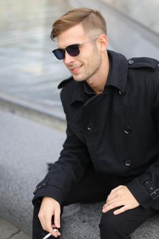 Young smiling man in sunglasses wearing black jacket sitting near glass Louvre Pyramid in Paris and smoking cigarette, France. Concept of male fashion model and urban photo session.