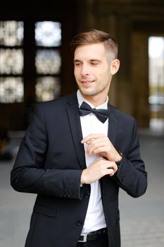 Young handsome man standing and posing, wearing black suit and bow tie, window in background. Concept of fashionable businessman and male model.