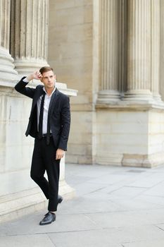 Young caucasian handsome man wearing black suit with tie leaning on column of building. Concept of groom and male fashionable model.