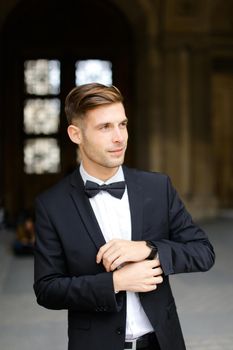 Young man standing and posing, wearing black suit and bow tie, window in background. Concept of fashionable businessman and male model.