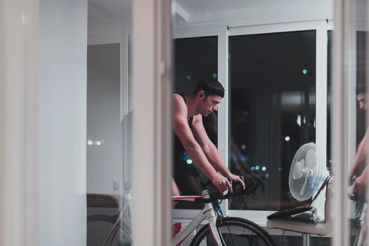 Man cycling on the machine trainer he is exercising in the home at night. Playing online bike racing game during coronavirus covid19 lockdown. New normal concept.